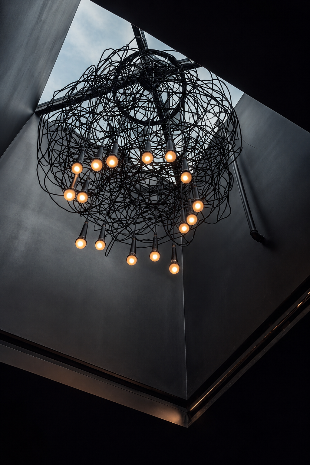 Close-up angle of the sculptural wire pendant light fixture at the Paul JAS Center Caption: Alternate angle of the Paul JAS Center's custom wire-and-bulb chandelier, showing the handmade, tactile detailing of the fixture against the skylight above.