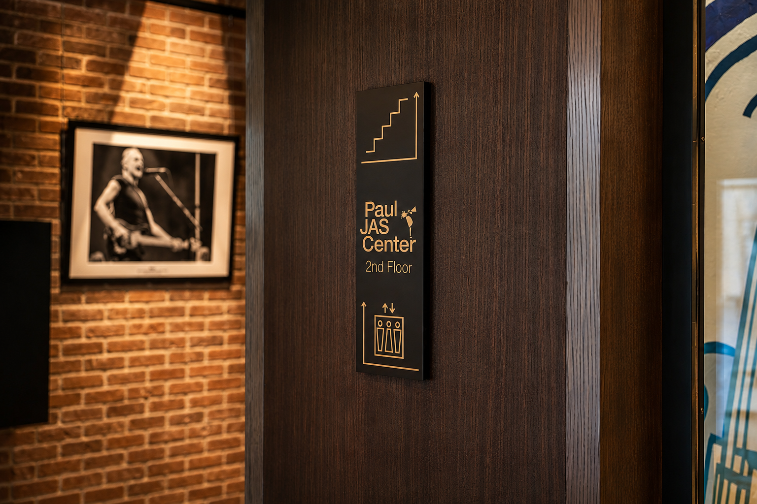 "Paul JAS Center 2nd Floor" wayfinding sign with stair and elevator icons on exposed brick Caption: Interior directional wayfinding signage at the Paul JAS Center, mounted on wood paneling beside an exposed brick wall, guiding guests to the second fl