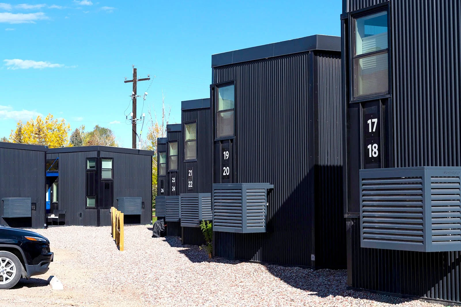 Row of black corrugated-metal tiny homes with numbered unit signage at Tiny Town

A row of black corrugated-metal tiny homes lines the gravel drive at Tiny Town, each pair of units identified with large numbered signage.