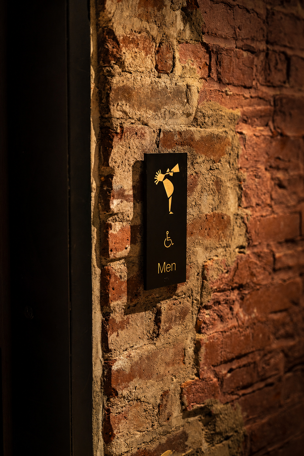 Men's restroom ADA sign with custom musician icon on exposed brick at the Paul JAS Center Caption: ADA-compliant restroom identification signage at the Paul JAS Center, featuring the venue's custom musician pictogram alongside the accessibility symbo