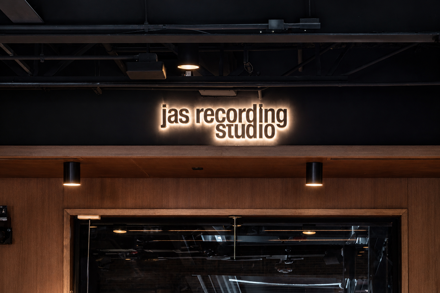 Backlit "JAS Recording Studio" sign above the studio's glass entry doors Caption: Custom backlit dimensional signage for the JAS Recording Studio inside the Paul JAS Center, mounted above the studio's glass entry doors on dark wood paneling.