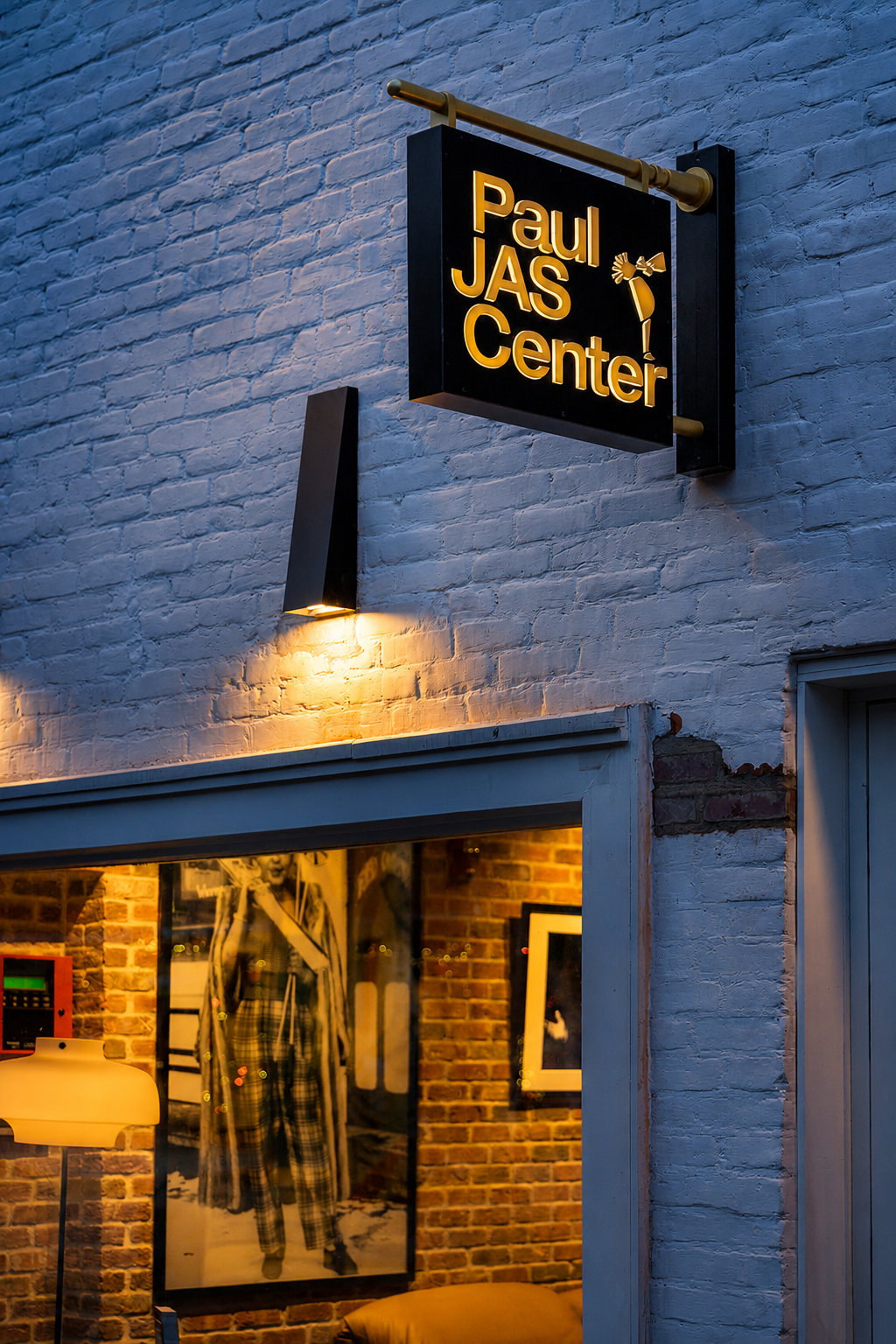 Close-up of the Paul JAS Center's perpendicular-mounted blade sign on brick at twilight Caption: Detail shot of the Paul JAS Center's exterior blade sign and matching wall sconce on painted brick, illustrating the brand's custom typography and musici