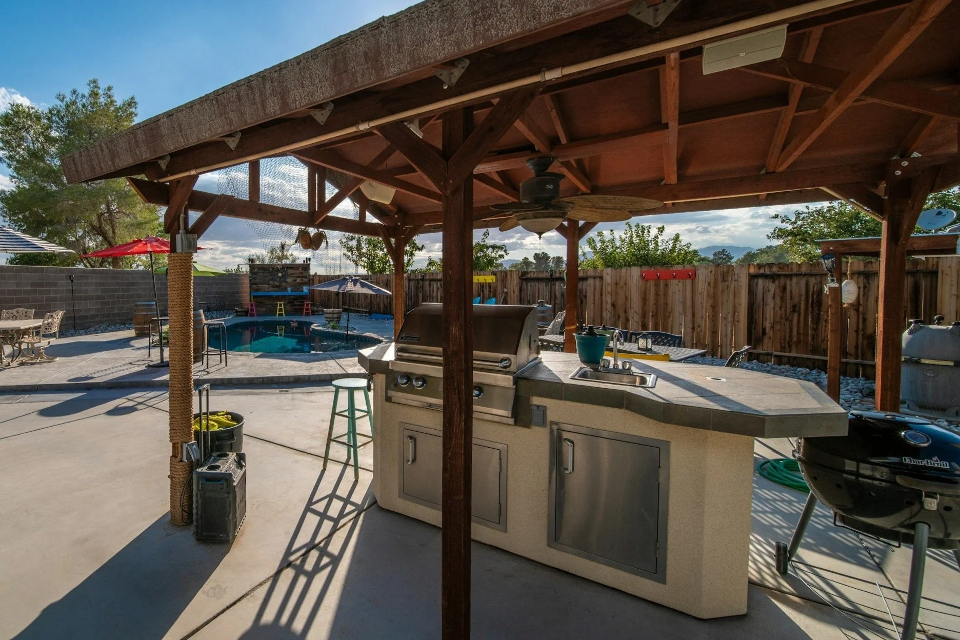 Outdoor patio with a covered barbecue area and swimming pool surrounded by a wooden fence, patio furniture, and umbrellas.