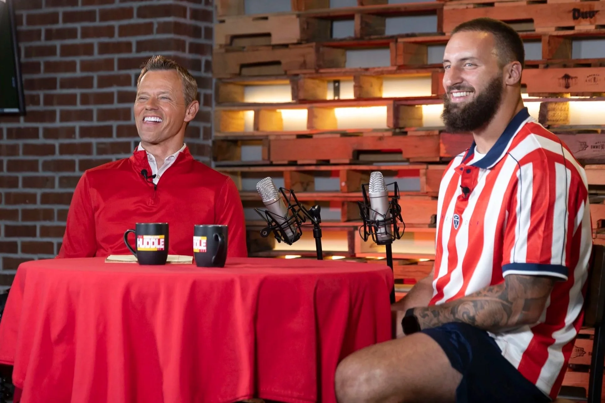 Two men sitting at a table with microphones, during a podcast or interview. The man on the left is wearing a red jacket, smiling, with a brick wall background. The man on the right has a beard, tattooed arm, and is wearing a red, white, and blue striped shirt, also smiling.