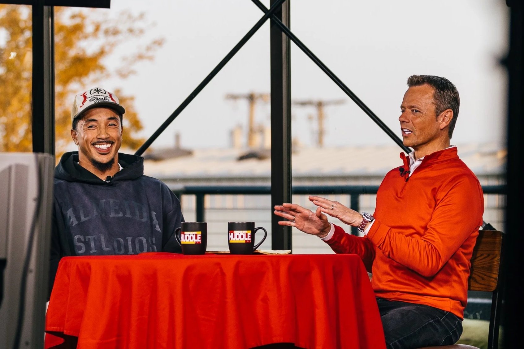Two men sitting at a table covered with a red cloth, having a conversation. One is smiling, wearing a hoodie and cap, the other in an orange jacket. There are two mugs with a logo on the table.