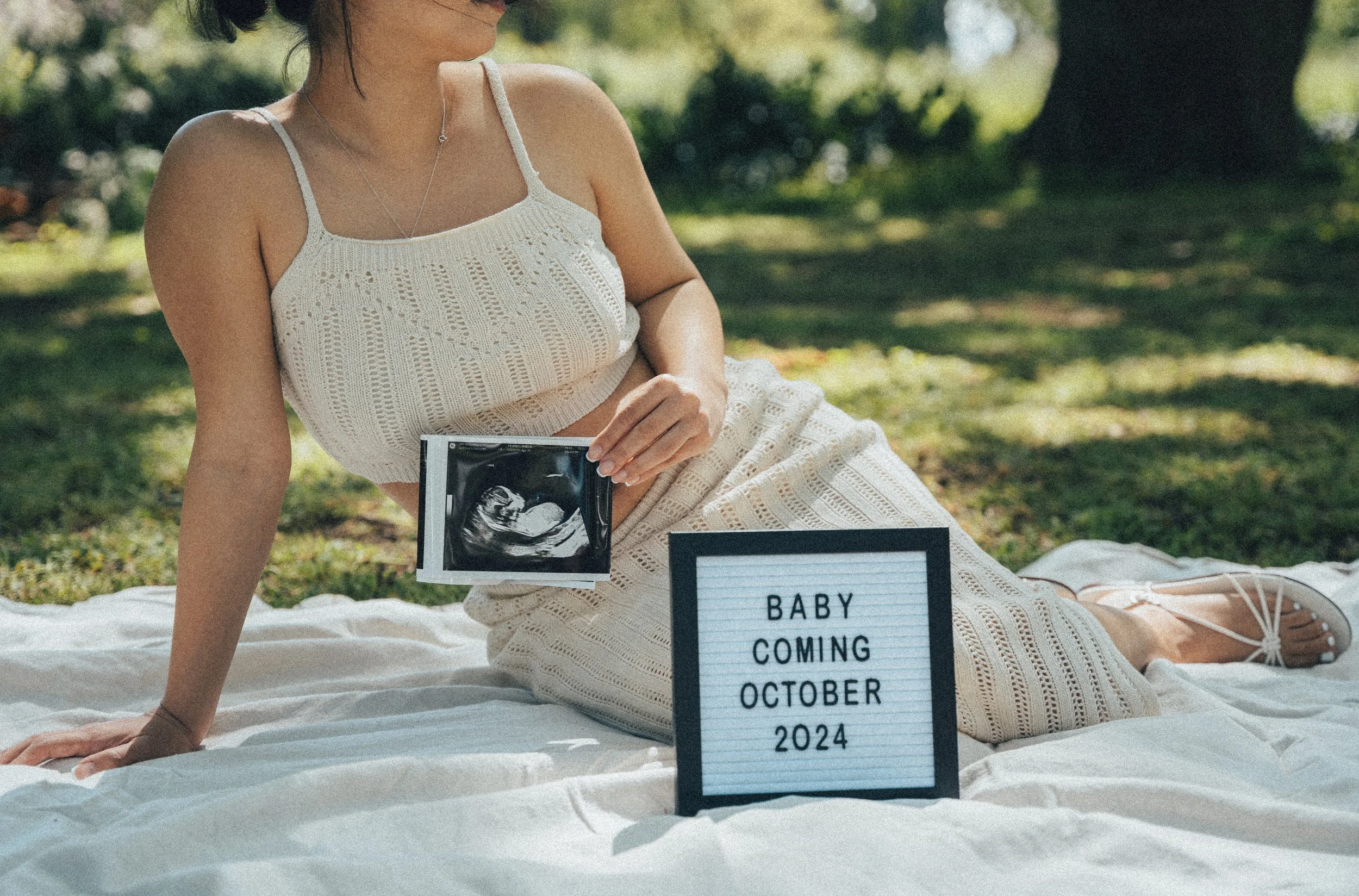 A woman in a white crocheted dress sitting outdoors on a blanket with a sign that reads "Baby Coming October 2024." She is holding a sonogram picture. The background is a grassy, shaded area with trees.