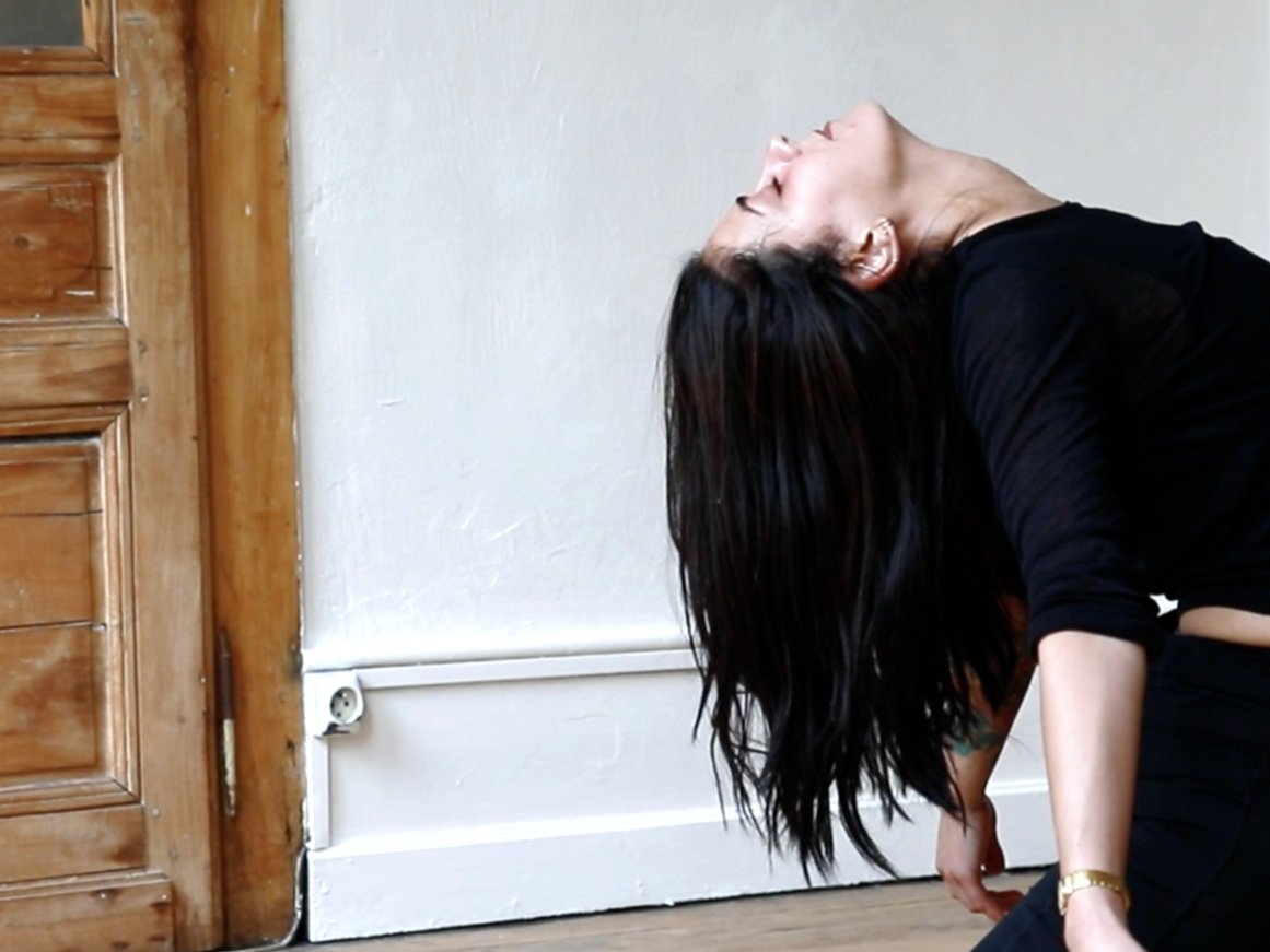 Femme en position de yoga ou de méditation, dos arqué, cheveux noirs longs, portant une robe noire, dans une pièce avec porte en bois et mur blanc
