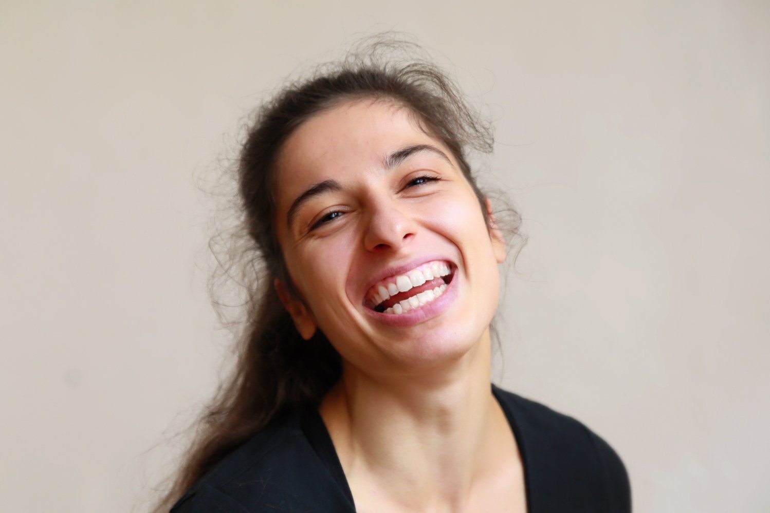 Une femme souriante avec des cheveux bruns, portant un t-shirt noir, en gros plan contre un mur beige.
