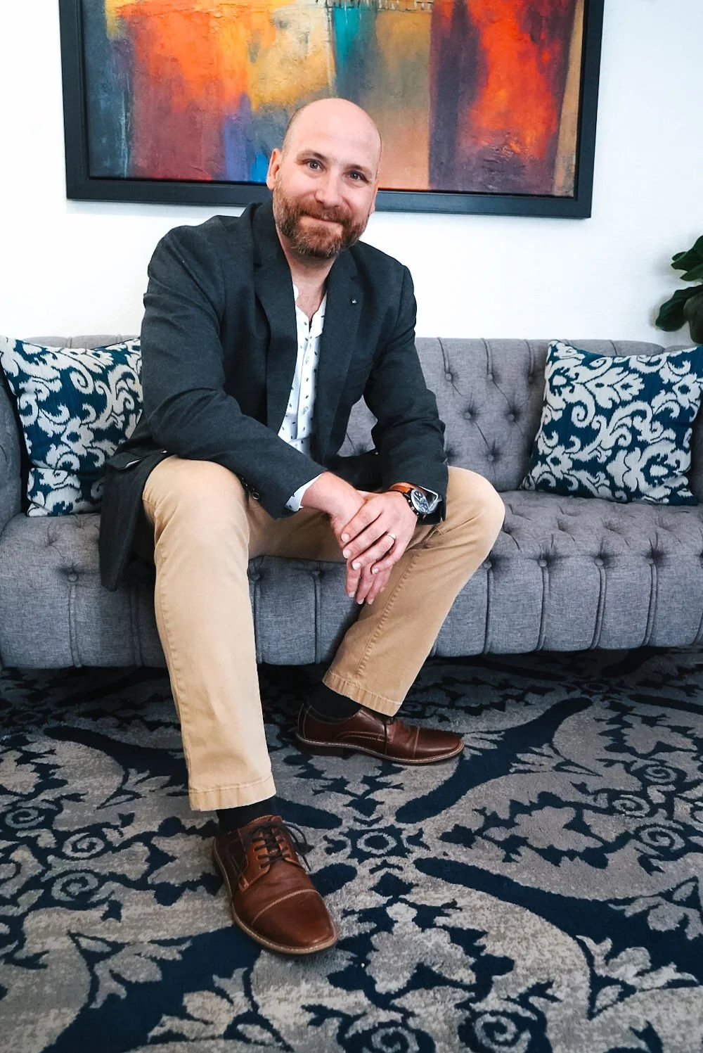 Man sitting on gray tufted sofa with decorative pillows, wearing a blazer and beige pants, colorful abstract painting in background.
