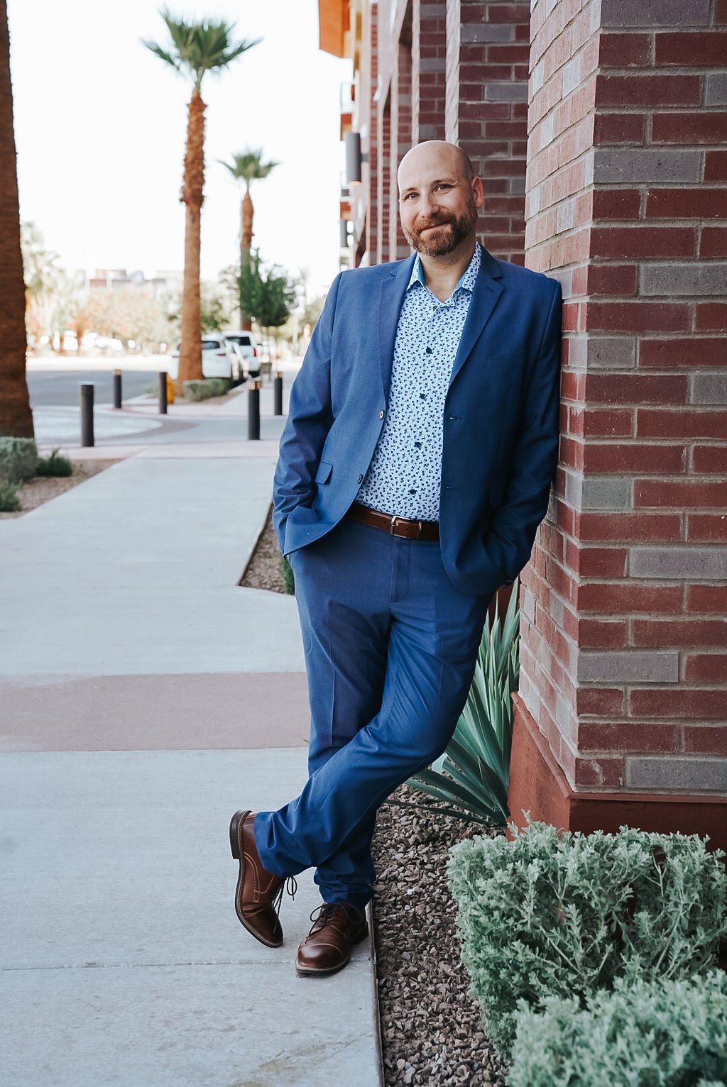 Man in blue suit leaning against brick wall on urban sidewalk with palm trees.