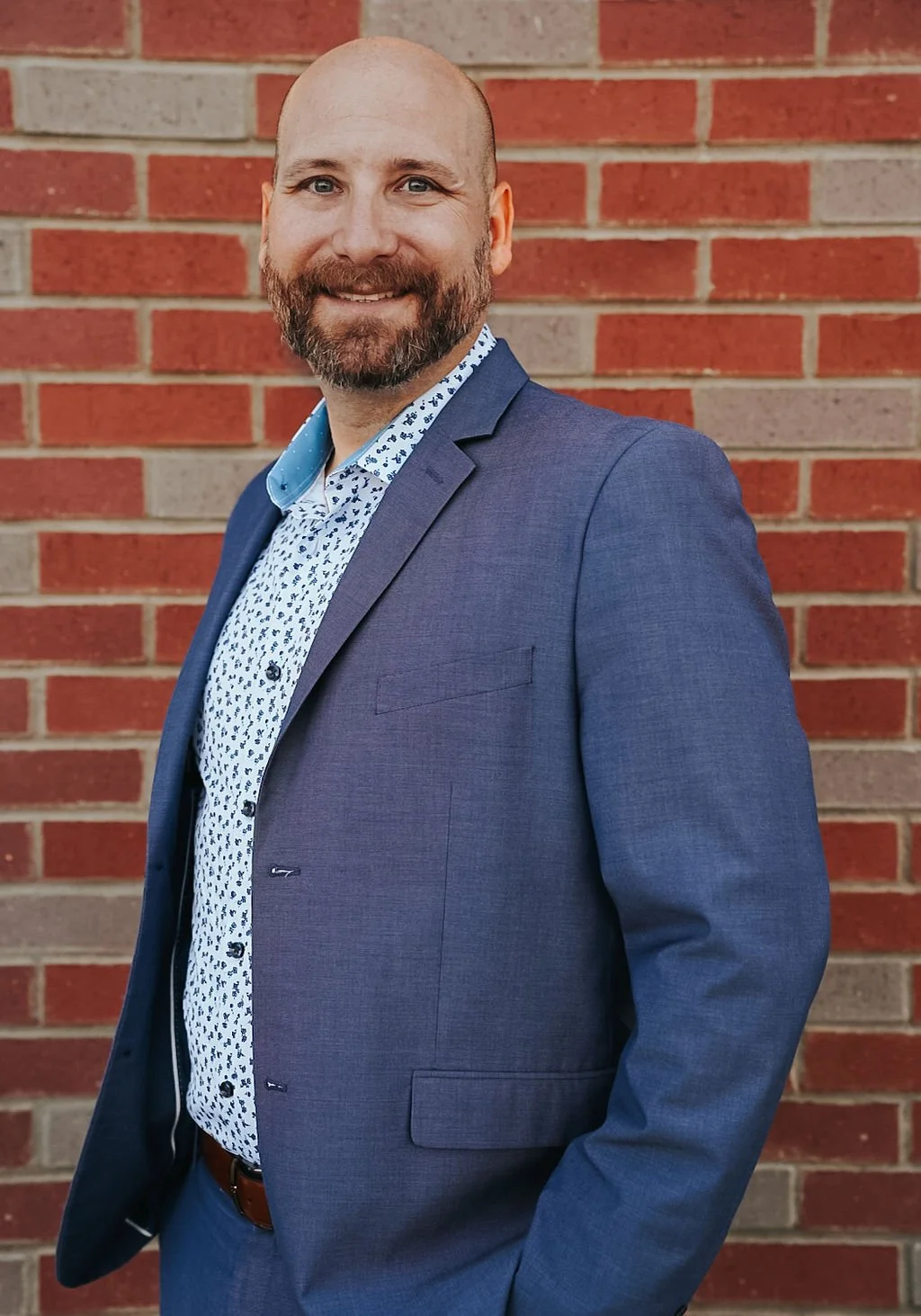A man in a blue suit with a patterned white shirt, standing in front of a red brick wall, smiling at the camera.
