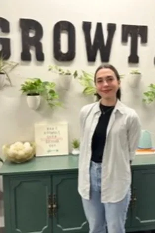 A woman standing in front of a wall with black letters spelling 'GROWTH'. She is smiling, wearing a white shirt over a black top, and light blue jeans. There are potted plants and a bowl on a green cabinet behind her.