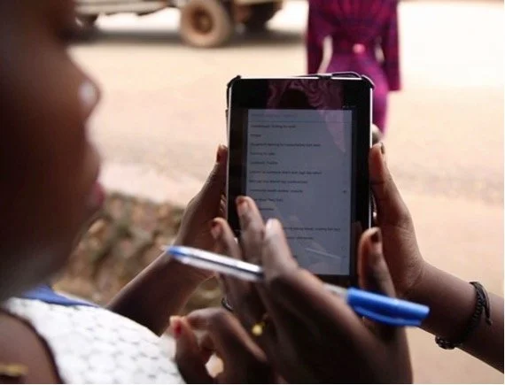 A person holding a smartphone and a pen, looking at the screen, with a blurred background showing another person and a vehicle.