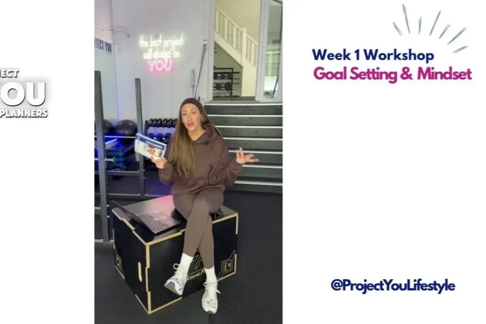 A woman sitting on a black box in a gym, holding a tablet, with a laptop nearby, during a workshop on goal setting and mindset.