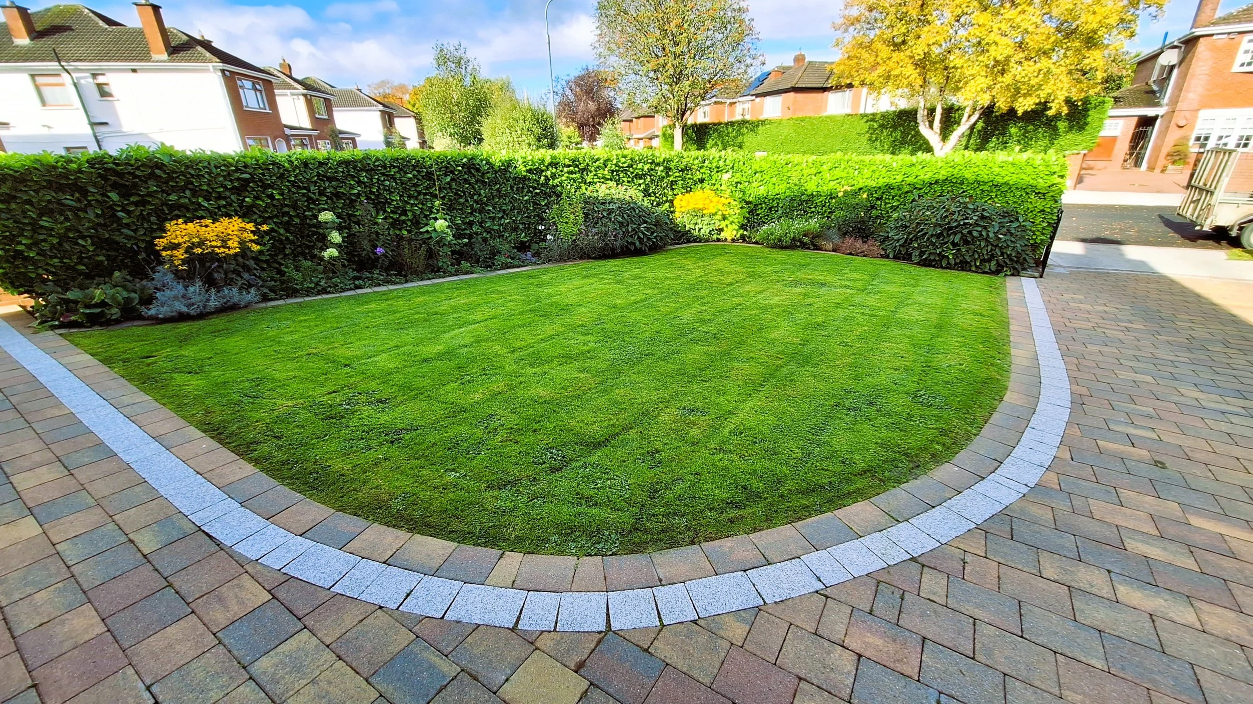 Well-maintained front yard with a lush green lawn, bordered by colorful flower beds, surrounded by a paved driveway and tall hedges, residential houses in the background, clear sky.