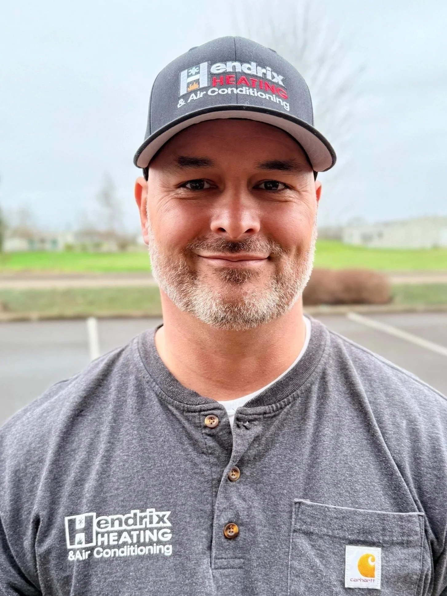 Man in a blue shirt and cap standing outdoors with trees and blurred background.