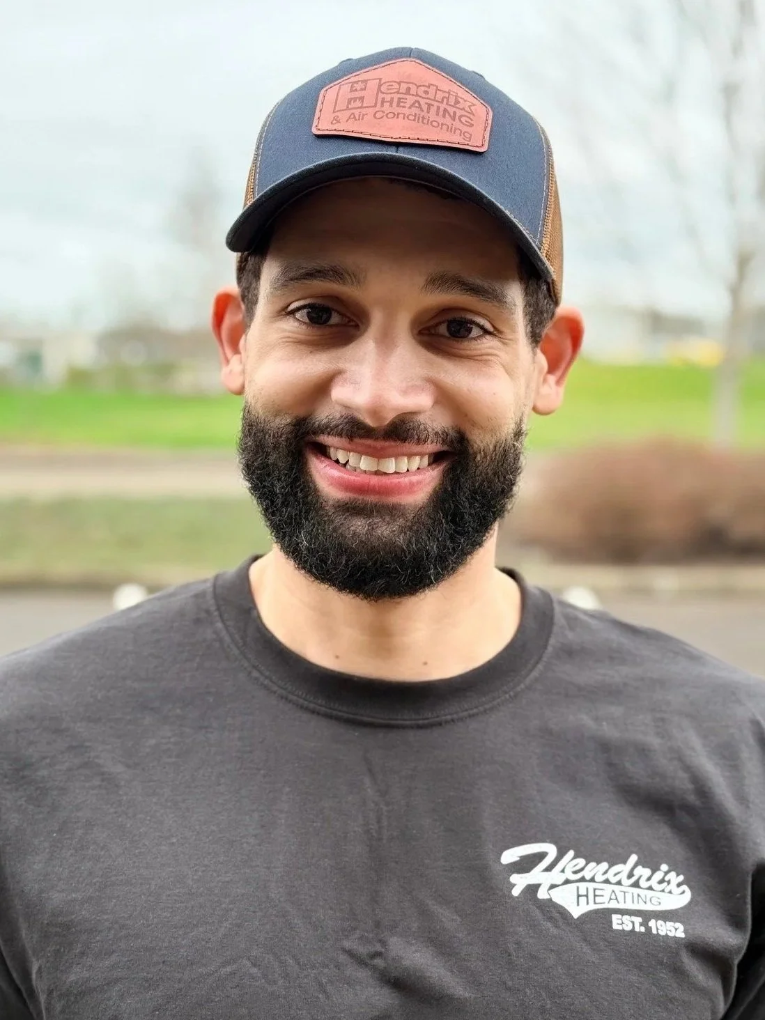Man wearing a "Hendrix Heating & Air Conditioning" cap and hoodie, standing outdoors with blurred trees in the background.