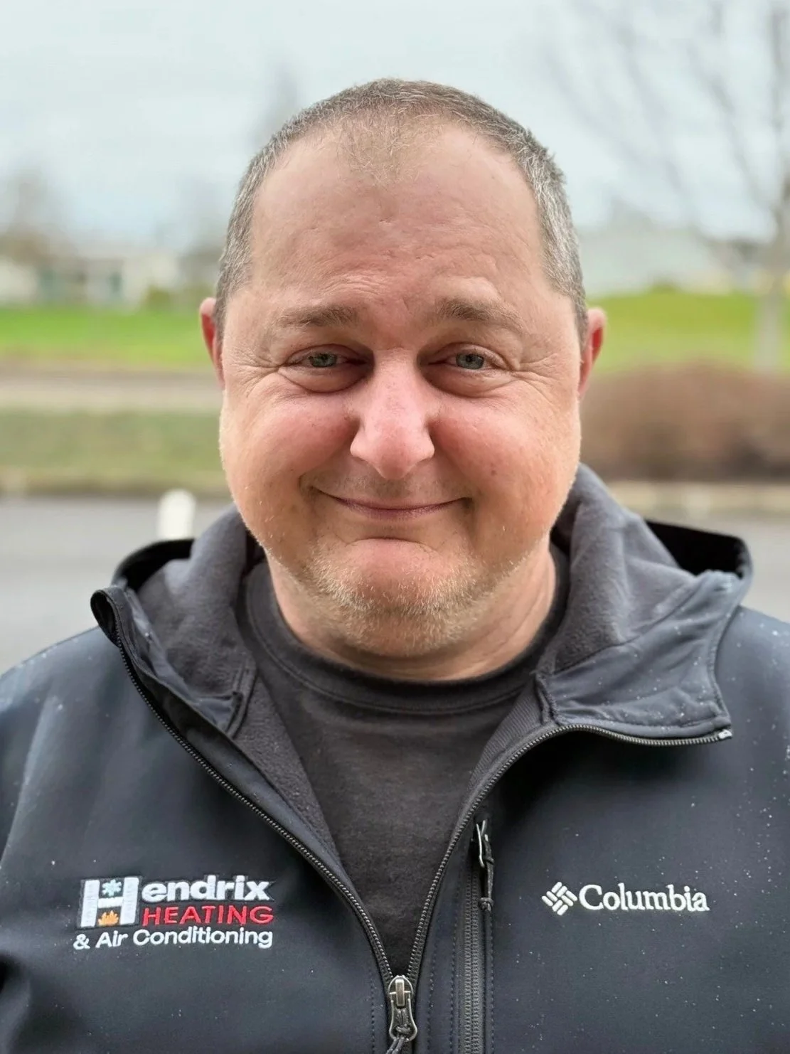 Person wearing a black cap with logo and navy blue jacket in outdoor setting with blurred foliage background.