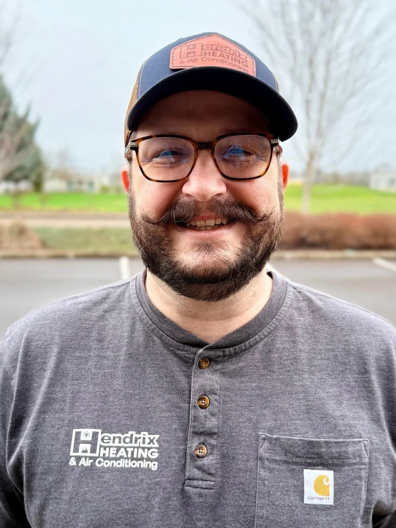 Man wearing a "Hendrix Heating & Air Conditioning" cap and shirt, outdoors with trees in the background.