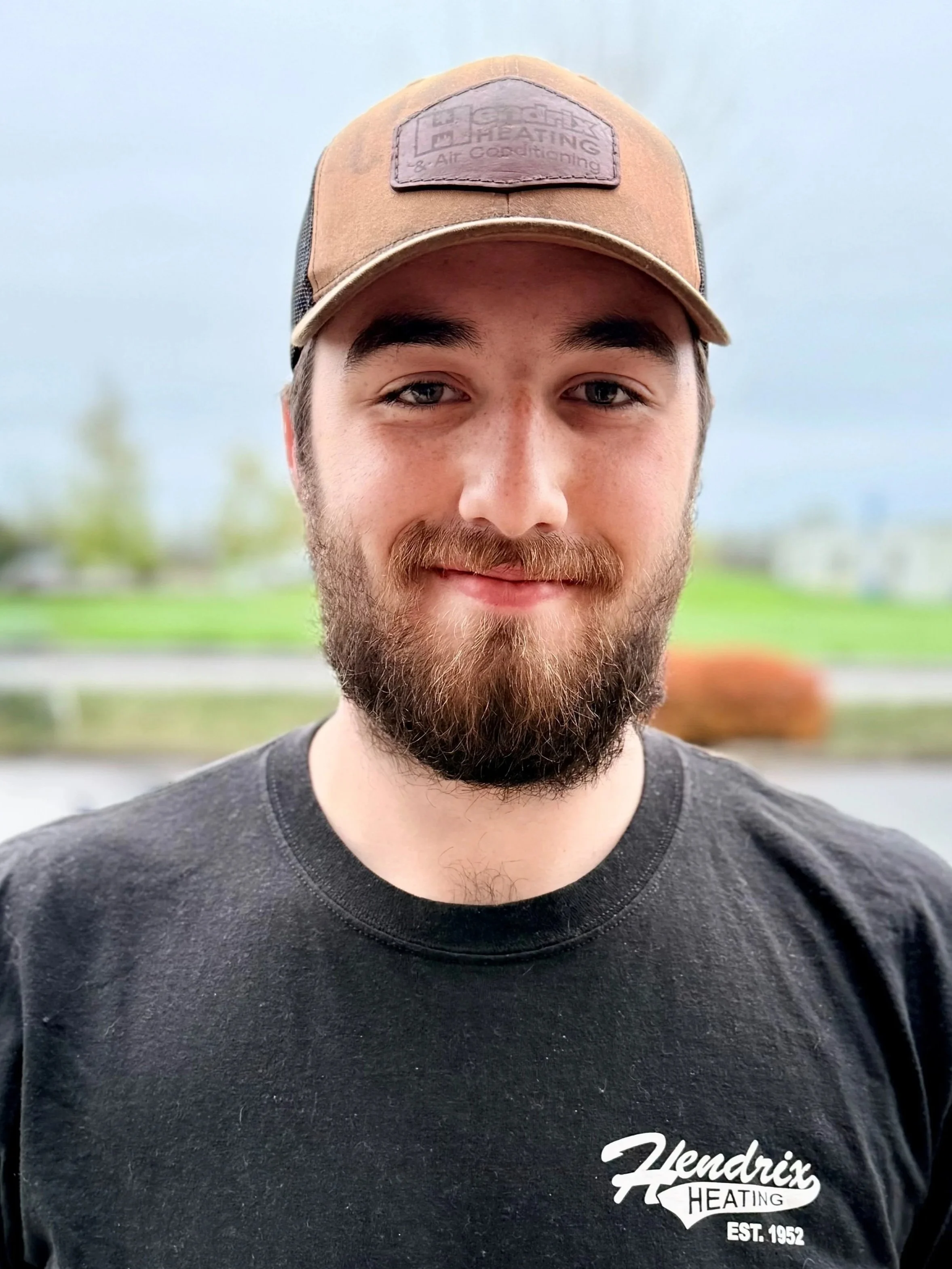 Man with a beard wearing a camo hat and a jacket with "Hendrix Heating" logo, standing outdoors with autumn foliage in the background.