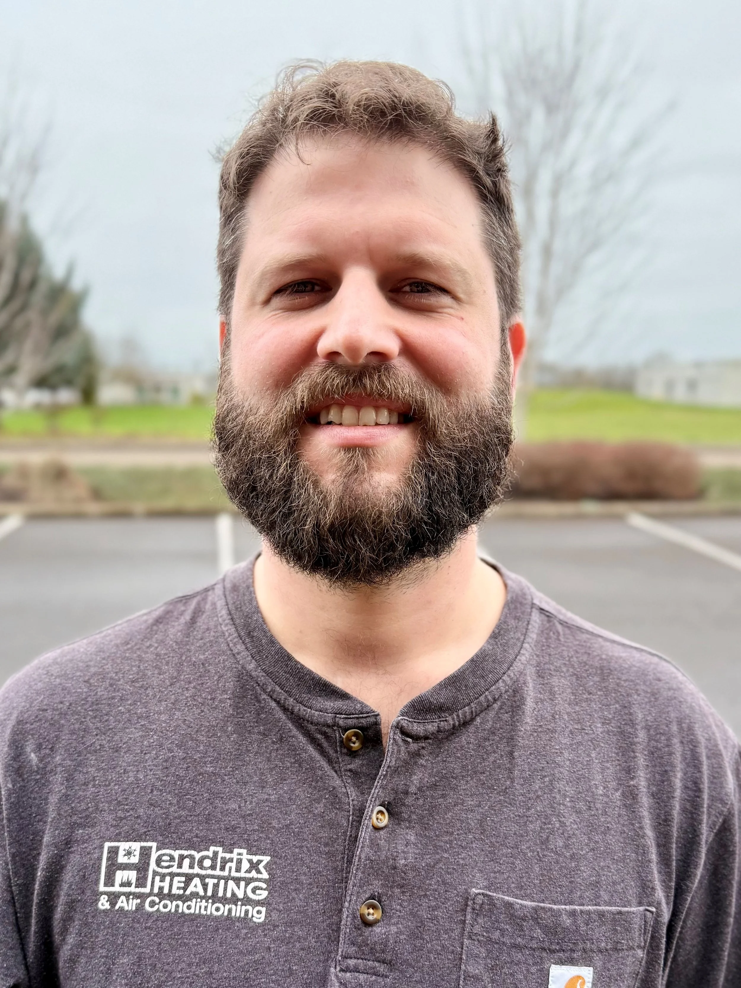 Smiling man with beard, wearing a black shirt, standing outdoors with trees and greenery in the background.