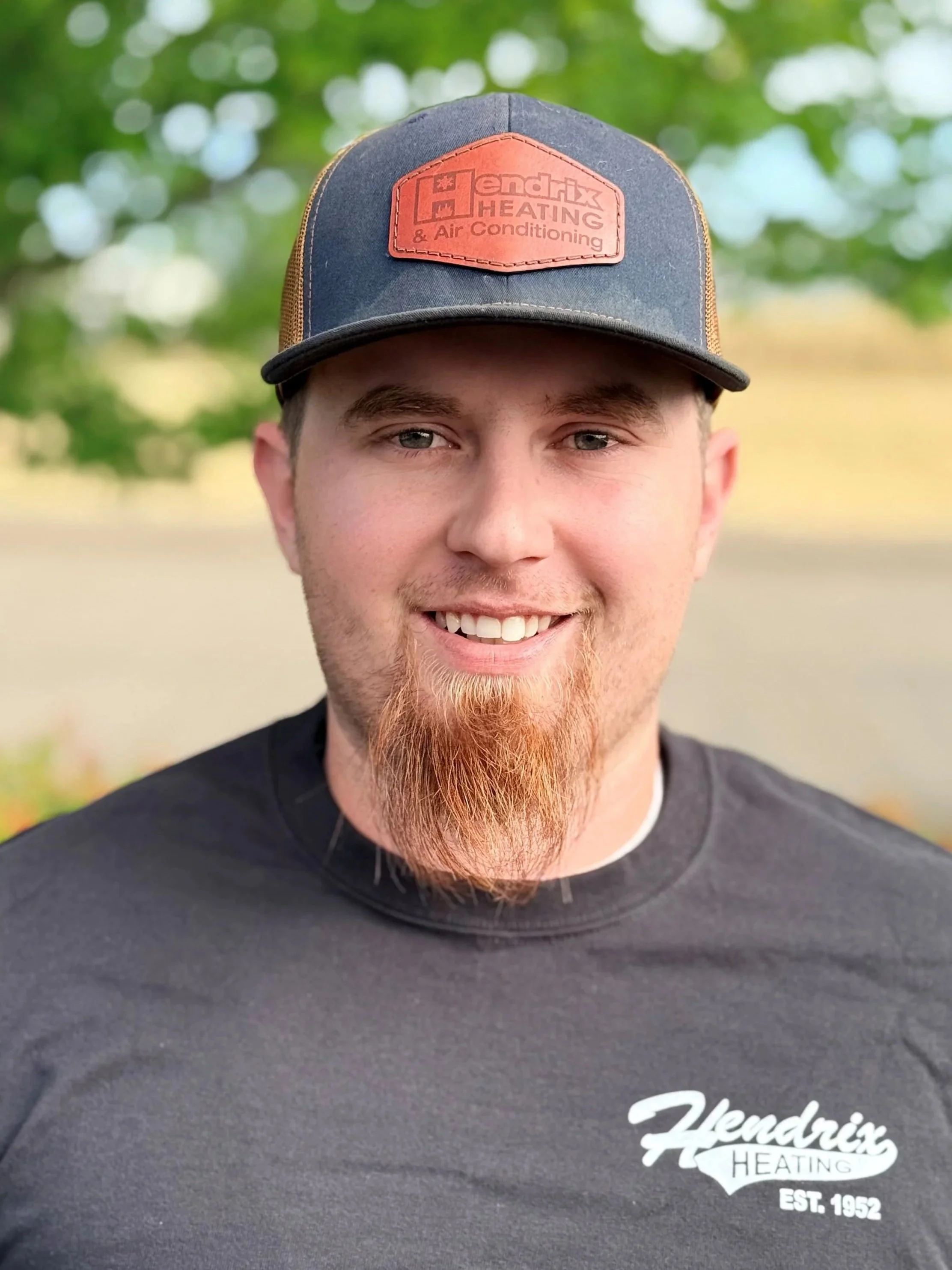 Close-up of a smiling man wearing a black baseball cap with a red patch that reads 'Hendrix Heating & Air Conditioning'. He is also wearing a black shirt with white text that reads 'Hendrix Heating Est. 1952'. The background features out-of-focus green trees.