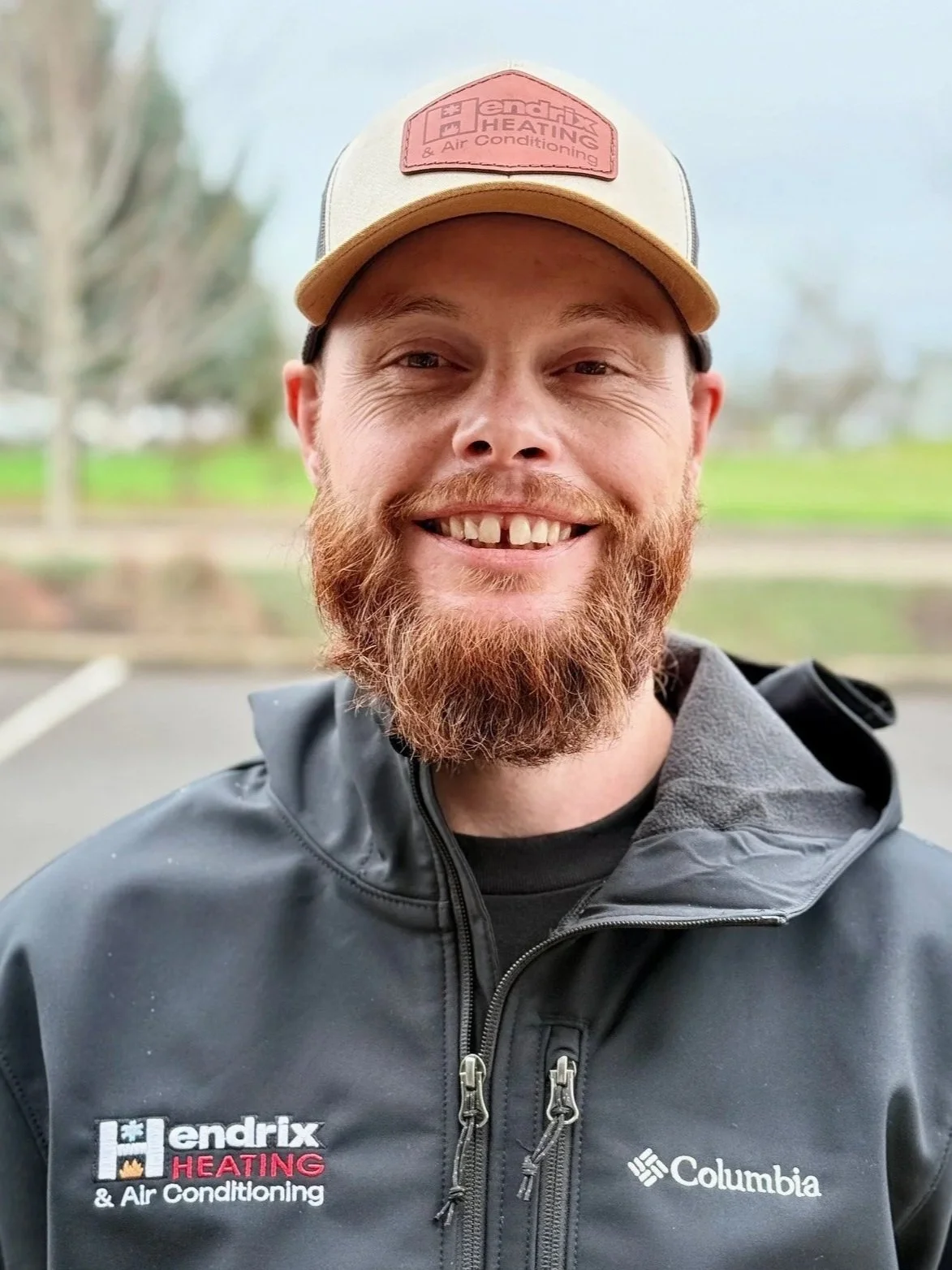 Man smiling outdoors wearing sunglasses and a black shirt with "Hendrix Heating" logo, with a blurred natural background.