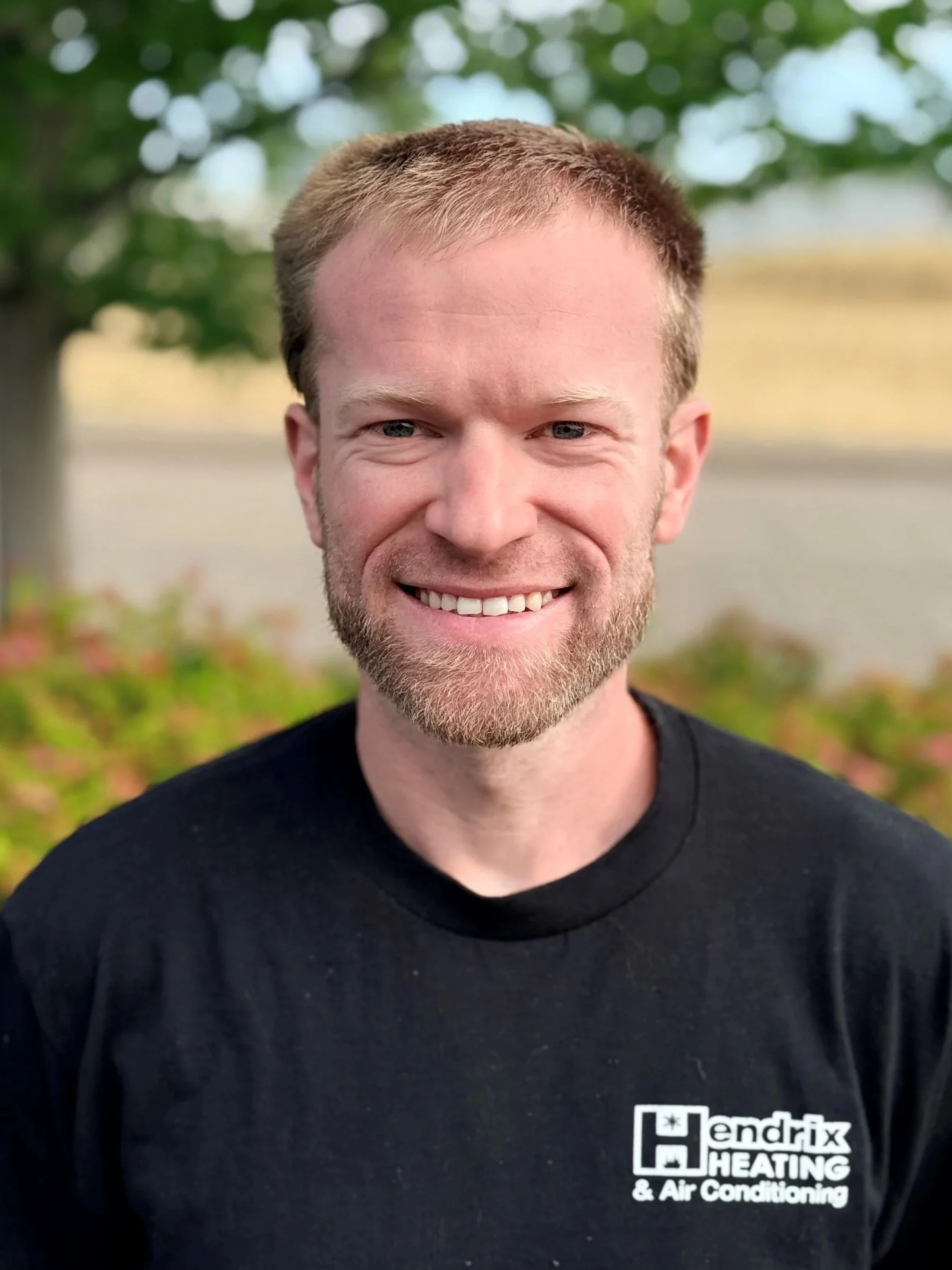 Close-up of a smiling man with short light brown hair and a beard, wearing a black Hendricks Heating & Air Conditioning shirt, standing outdoors with blurred green trees and a body of water in the background.