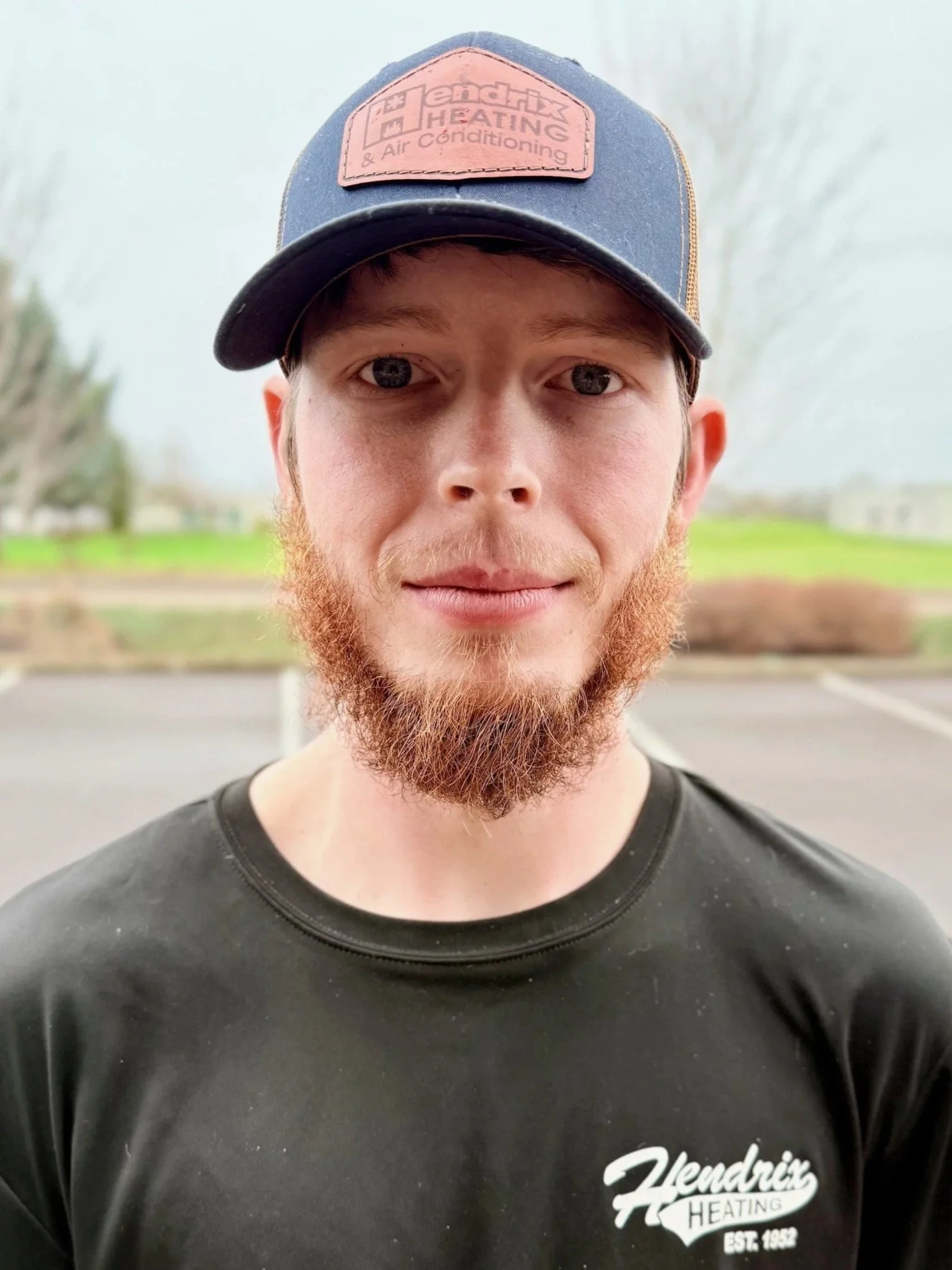 Smiling man wearing a Hendrix Heating hoodie and cap, standing outdoors with blurred fall foliage in the background.