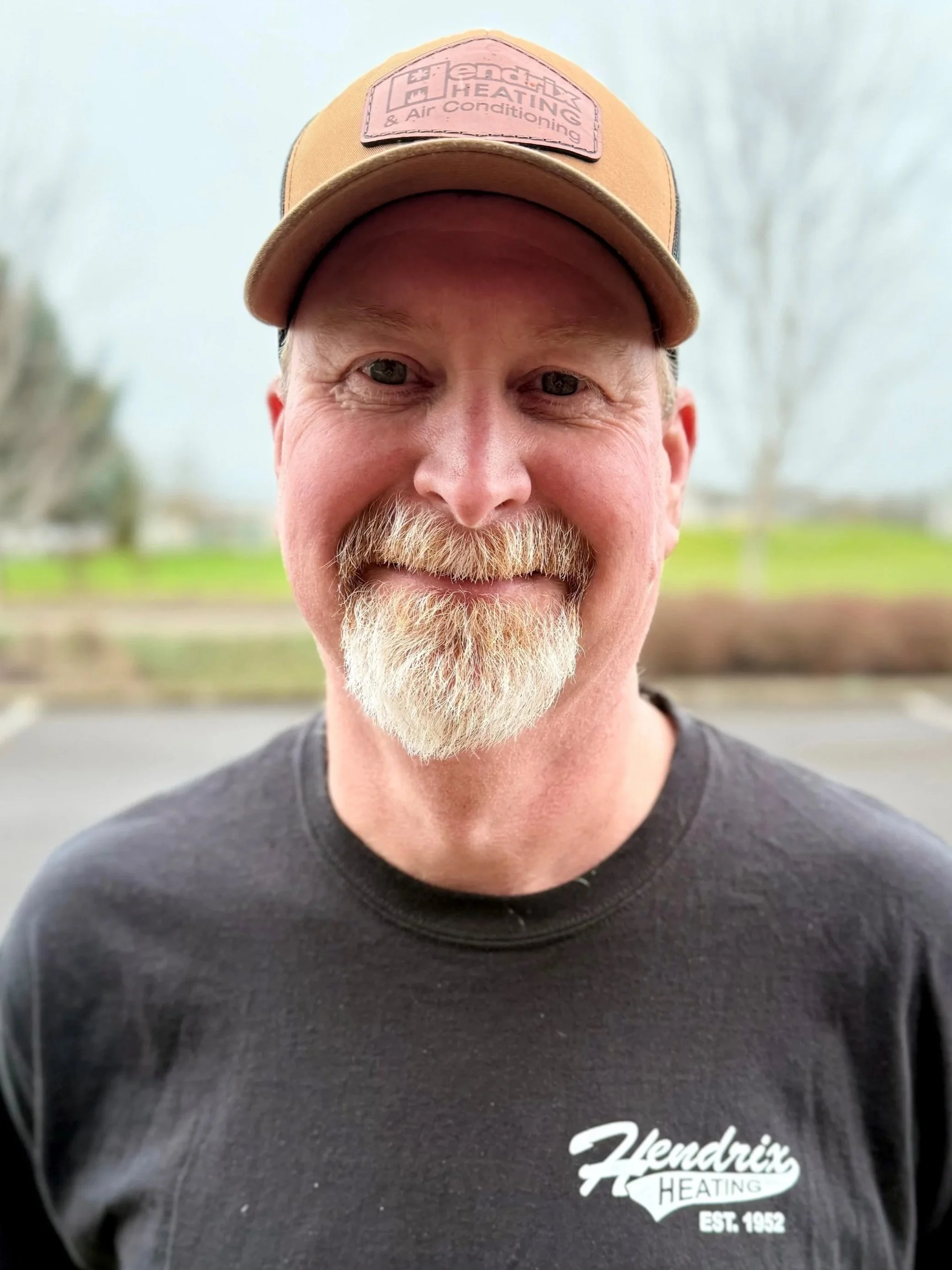 Man wearing a cap and jacket with "Hendrix Heating & Air Conditioning" logo, standing outdoors with trees in the background.