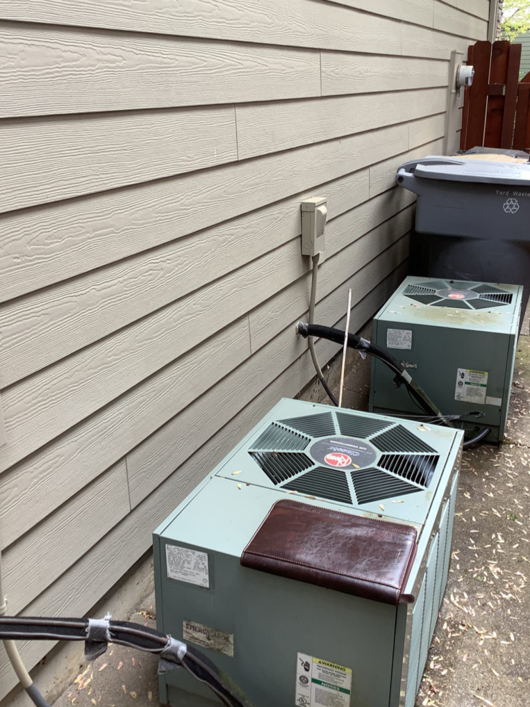 Before photo of two older top-discharge AC condensers in a tight side yard along the home’s exterior wall.