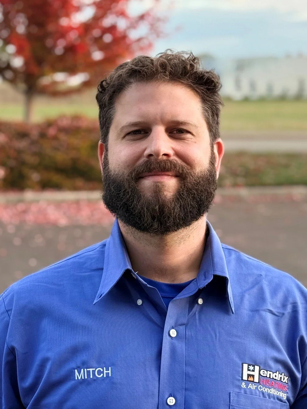 Smiling man with beard, wearing a black shirt, standing outdoors with trees and greenery in the background.