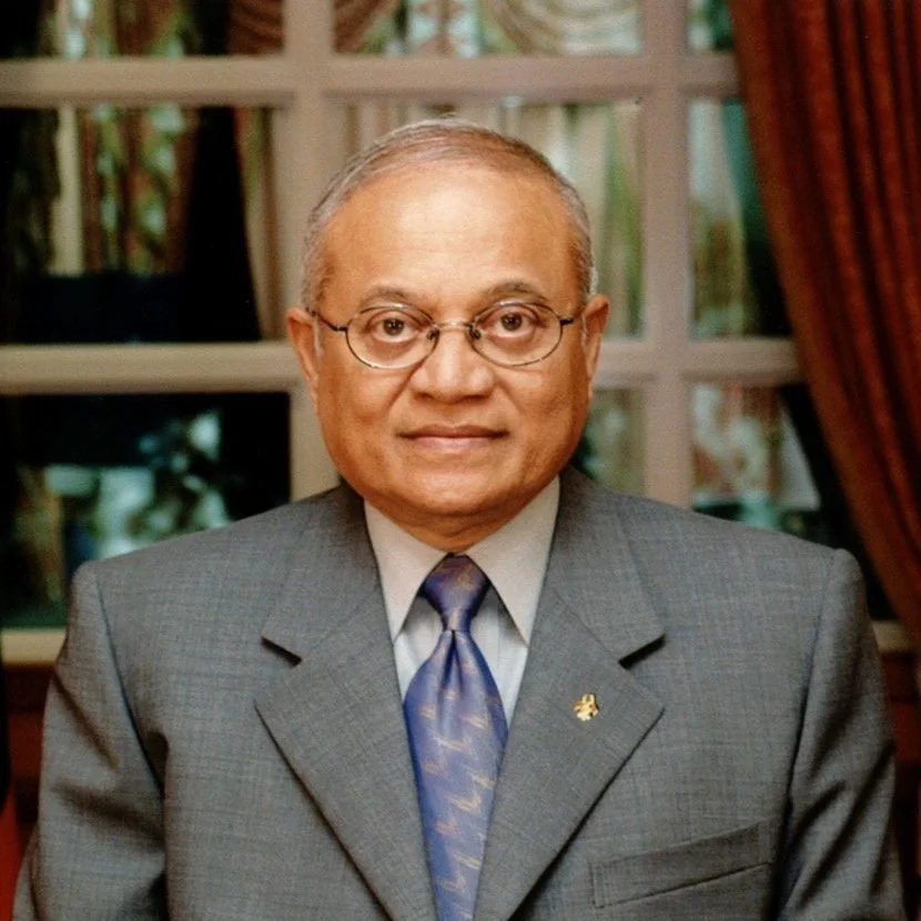 Maumoon n wearing glasses and a suit, standing in front of windows with curtains in a formal indoor setting.