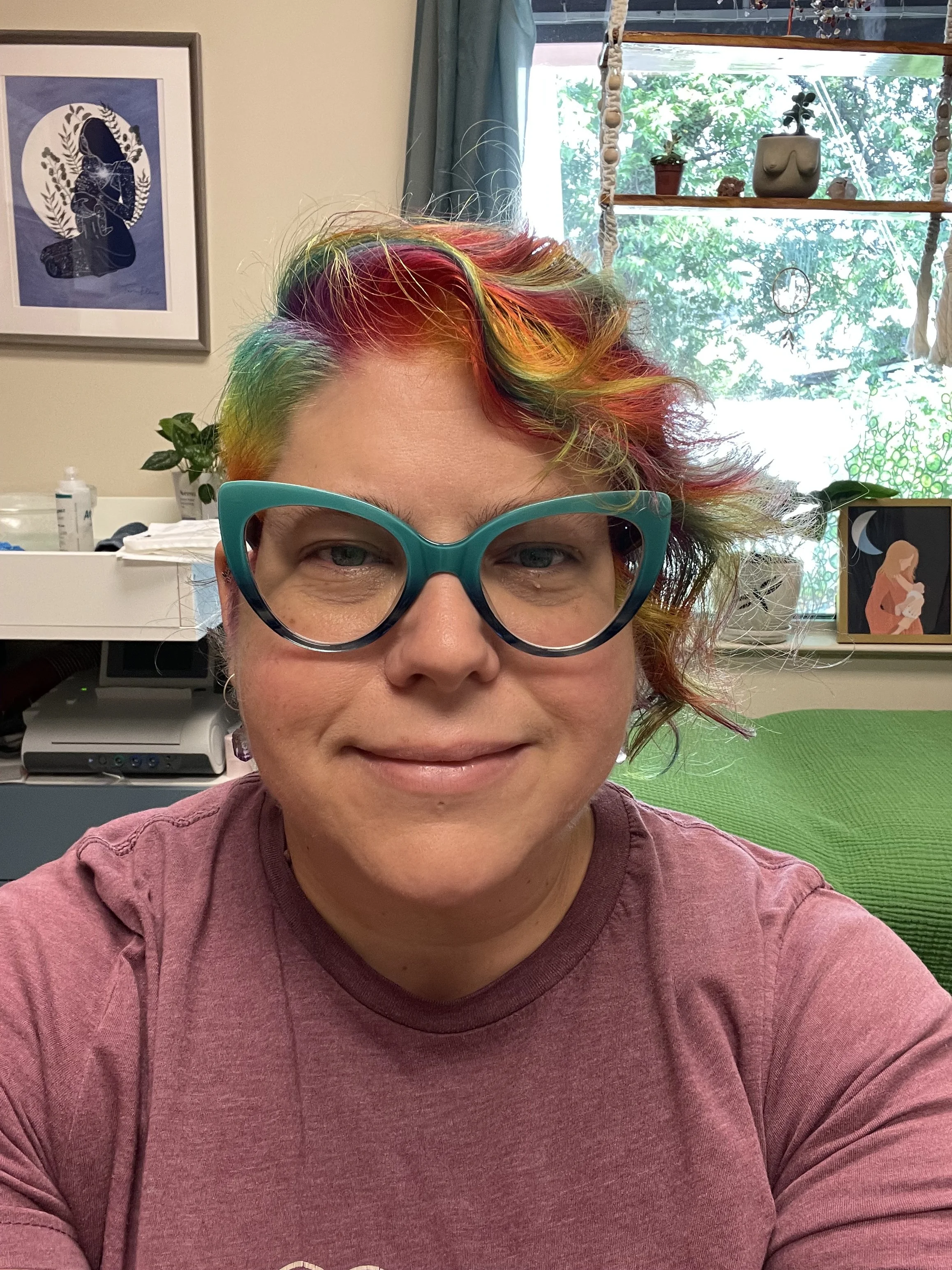 Headshot of the Midwife, Meagan.  Meagan is wearing teal green cat-eye glasses, a dusty rose colored shirt, and is smiling.