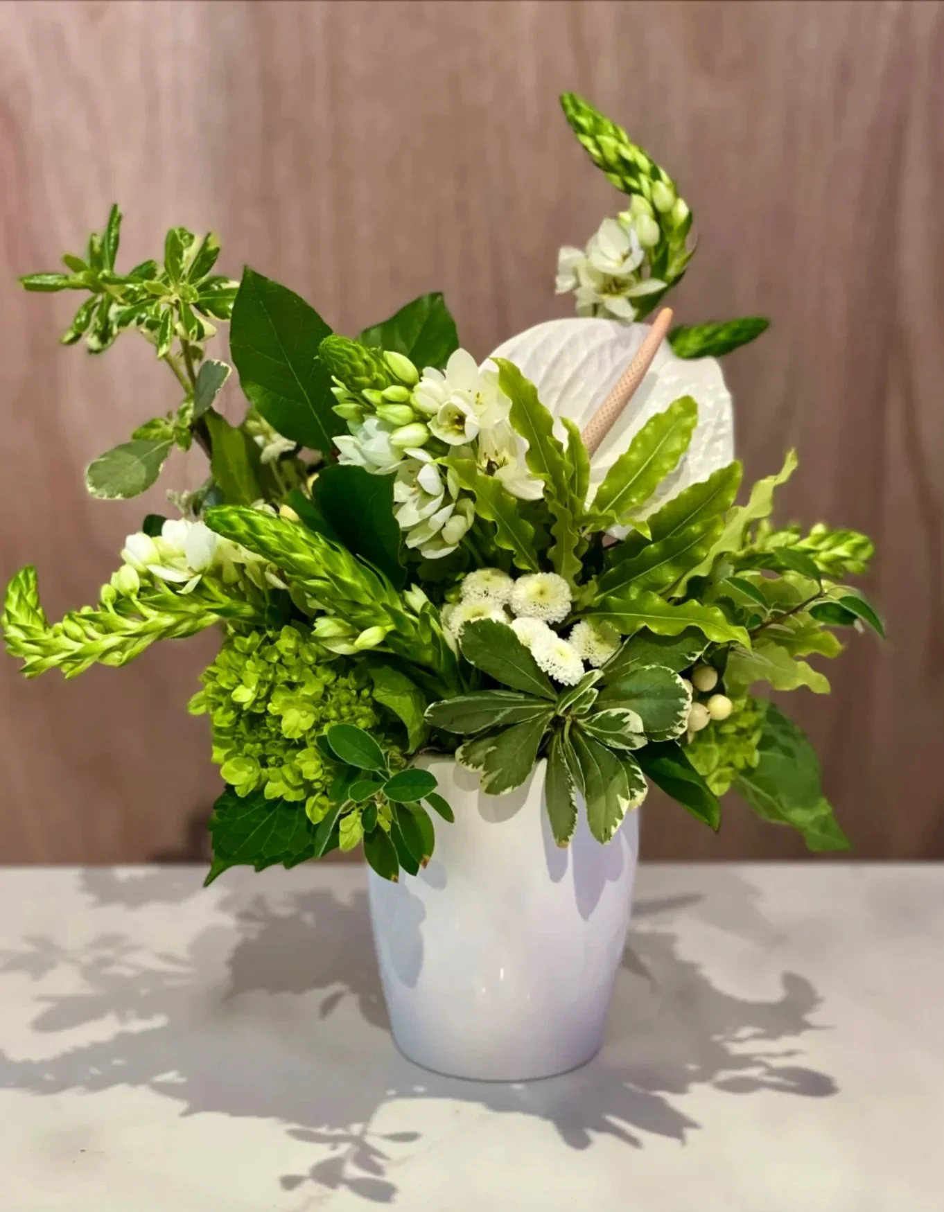 "Green and white floral arrangement in a white vase, featuring assorted leaves, blooms, and textured greenery."