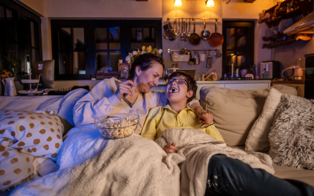 Two people sitting on a couch under blankets, watching something and sharing popcorn.