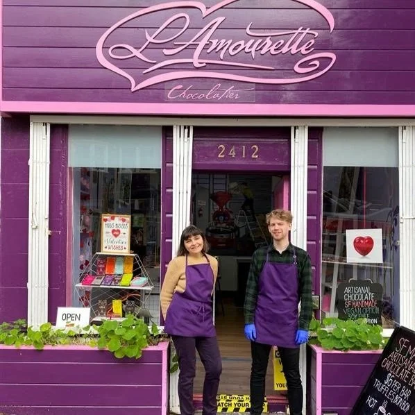 Two workers standing outside a purple storefront labeled “L’Amourette Chocolatier.”