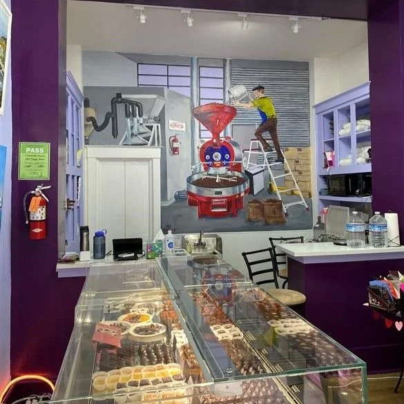 Interior of a chocolate shop with a glass display case and a mural of a person operating cocoa‑processing equipment.