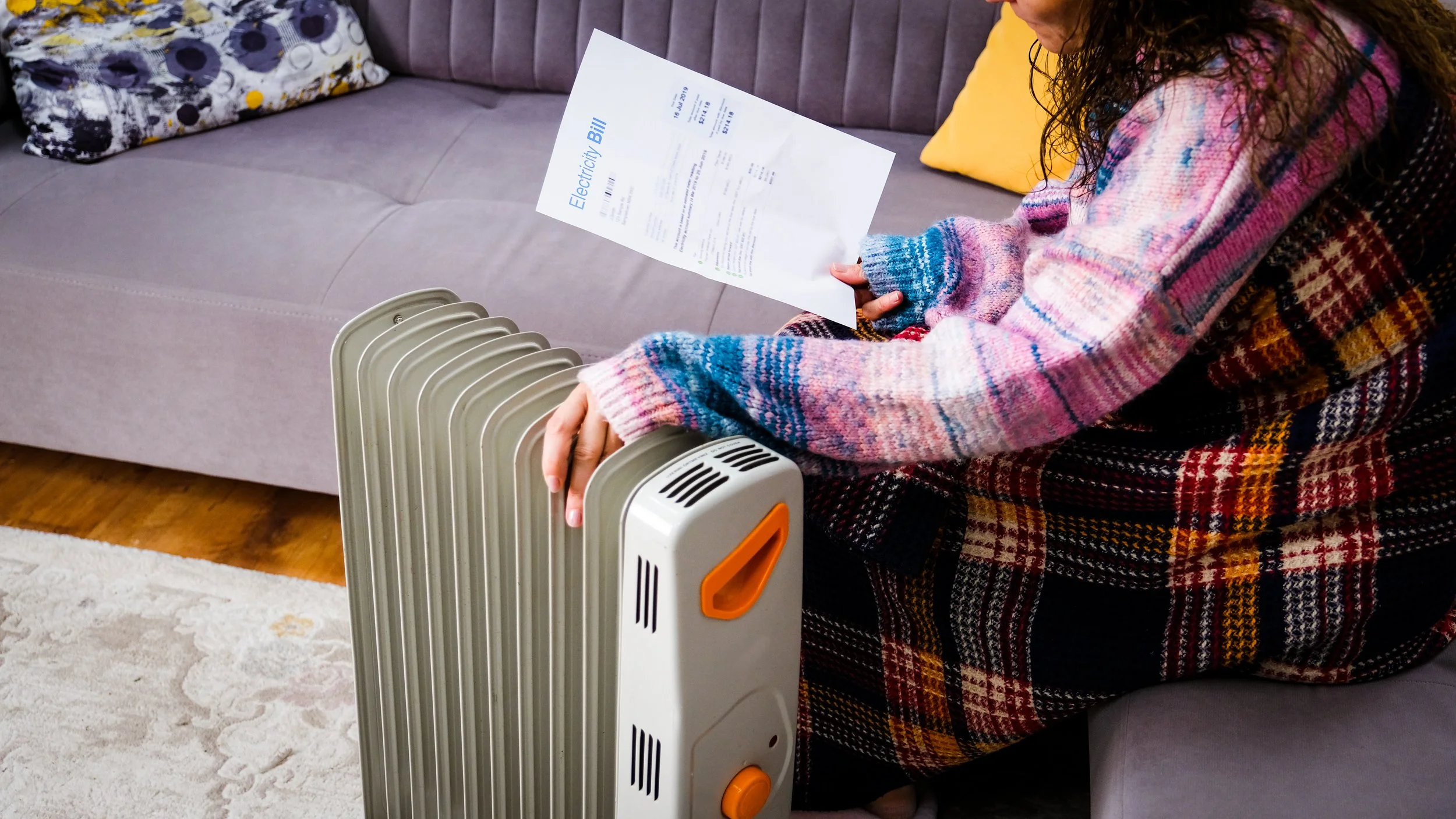 Person sitting on a couch holding an energy bill next to a portable space heater.