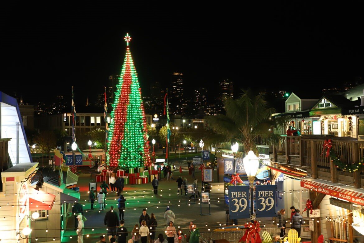 A large Christmas tree with red and green lights stands in a lively, decorated outdoor plaza at night. People walk around, and Pier 39 signs and festive lights are visible throughout the scene.