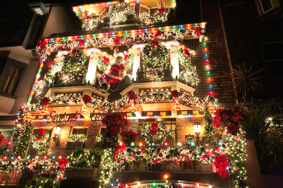 A house is decorated with colorful Christmas lights, wreaths, garlands, red bows, and a Santa Claus figure on the balcony, creating a festive holiday display at night.