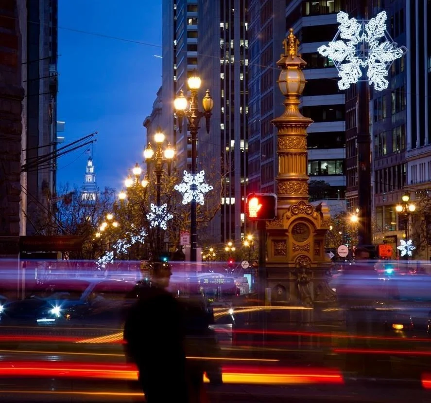 City street at dusk with tall buildings, decorated with illuminated snowflake lights. Streetlights glow, cars create light trails, and a blurred pedestrian stands in the foreground. A red pedestrian signal is visible.
