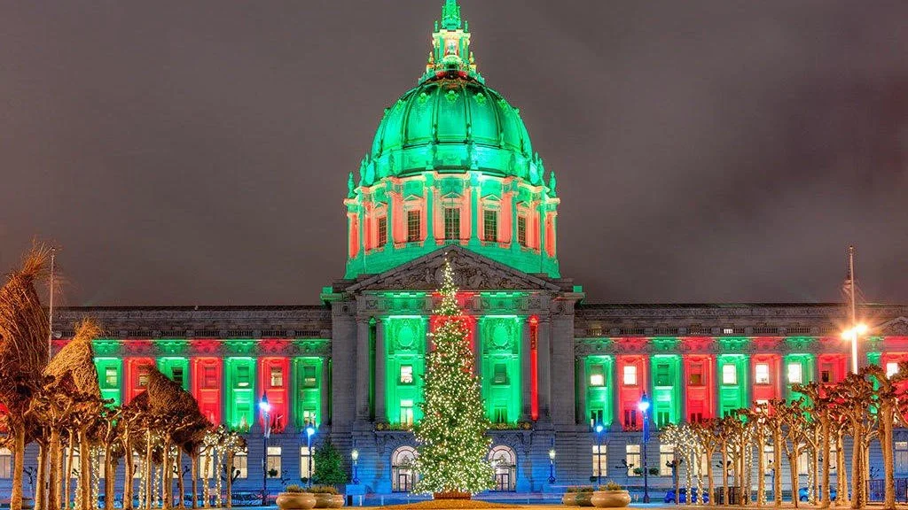 San Francisco City Hall is illuminated with green and red lights at night, with a decorated and lit Christmas tree in front and palm trees lining the walkway.