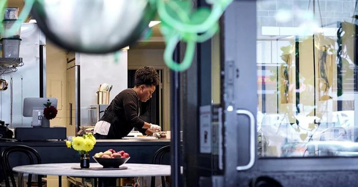A woman prepares food in a modern kitchen, seen through a glass door. A round table with flowers and a bowl of fruit is in the foreground, with kitchen utensils and decor partially visible.