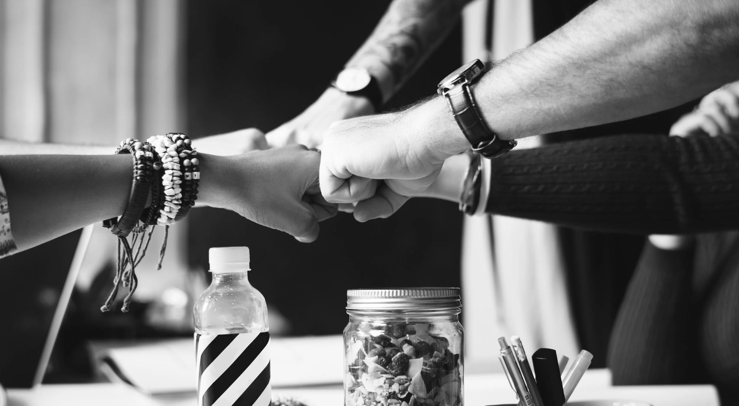 A black and white image shows four people bumping fists in a gesture of teamwork over a table with a striped water bottle, a jar of snacks, and pens.