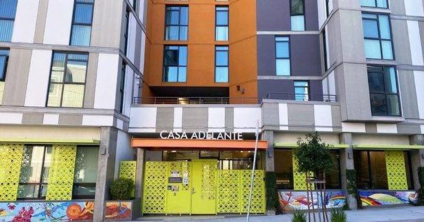 Modern apartment building with colorful orange and grey panels, large windows, a bright yellow entrance, and a sign reading Casa Adelante. Decorative panels and murals border the entrance area.