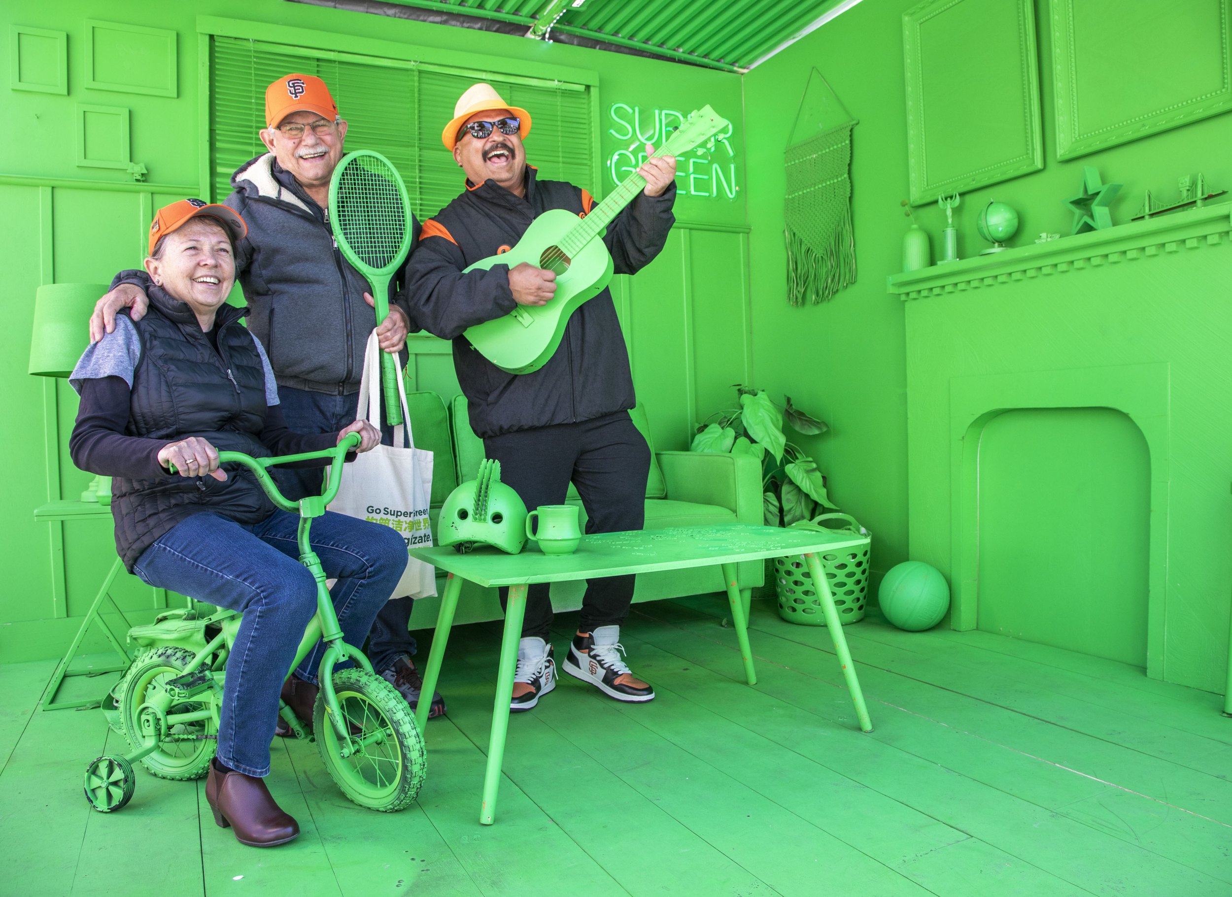 Three smiling adults pose in a room painted entirely green. One sits on a small green bike, another holds a tennis racket, and the third plays a green guitar.