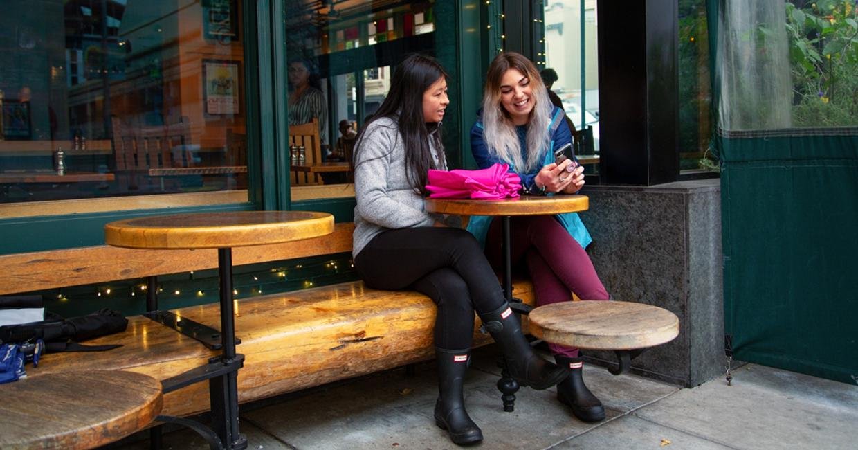 Two women sit at a wooden outdoor café table, smiling and looking at a phone together. One woman has dark hair and wears a gray jacket, the other has light hair and wears a purple jacket.