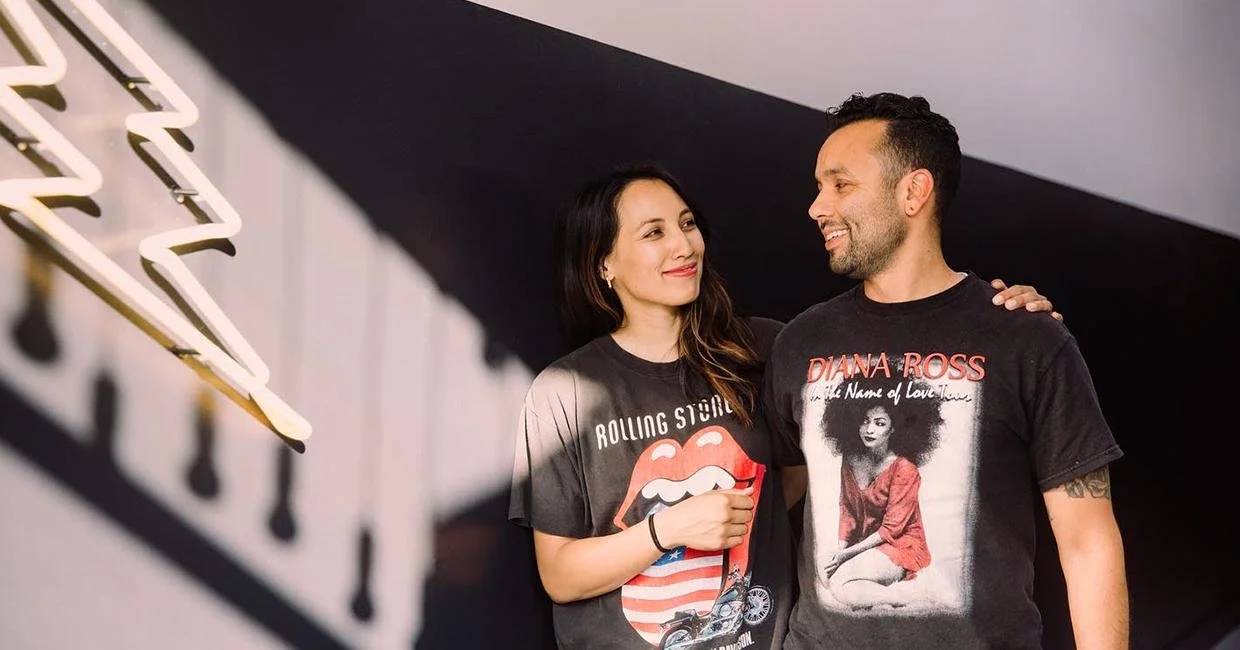 Two people standing together indoors, both wearing graphic T-shirts, with a shadowed wall and neon-style light in the background.