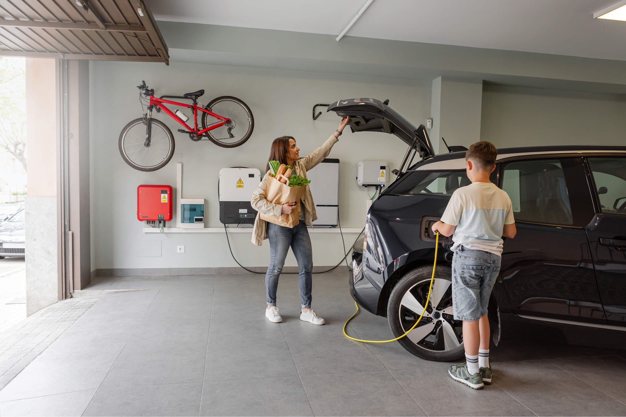 A parent and their child in their garage grabbing groceries from a charging electric vehicle.